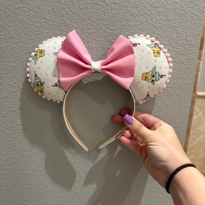 May include: Pink bow headband with white ears featuring a repeating pattern of a green character with a brown ice cream cone. The ears are trimmed with pink pom-poms.