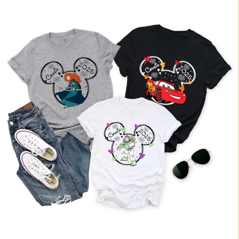 May include: Three graphic t-shirts with a Disney theme. One gray shirt features a cartoon image of Merida, the second is black with Lightning McQueen, and the third is white with Buzz Lightyear. Each shirt has "Cousin 2025" in a Mickey Mouse head design.
