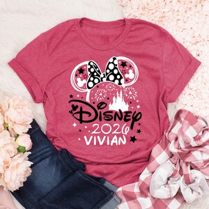 May include: A red t-shirt featuring a Minnie Mouse design with a polka dot bow, stars, and a castle. The text reads "Disney 2026 Vivian". The shirt is paired with blue jeans and a red and white plaid shirt.