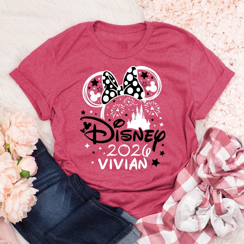 May include: A red t-shirt featuring a Minnie Mouse design with a polka dot bow, stars, and a castle. The text reads "Disney 2026 Vivian". The shirt is paired with blue jeans and a red and white plaid shirt.