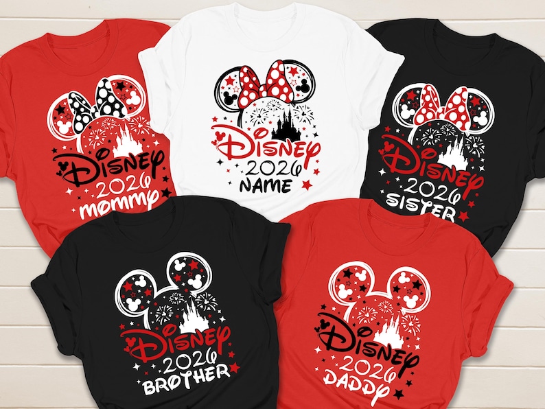 May include: A collection of Disney-themed t-shirts in red, white, and black. Each shirt features a Mickey Mouse head design with a bow, fireworks, and the text "Disney 2026" along with family member titles like "Mommy", "Sister", "Brother", and "Daddy".