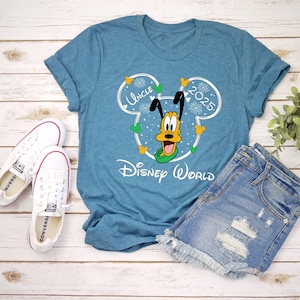 May include: A teal t-shirt with a graphic of Pluto in a Mickey Mouse outline, with the words "Uncle 2025" and "Disney World". The shirt is paired with denim shorts and white sneakers.