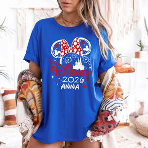 May include: Royal blue t-shirt featuring a Disney-themed graphic. The design includes Mickey Mouse ears, a bow, fireworks, and the text "Disney 2026 Anna" in red and white. The shirt has rolled-up sleeves.