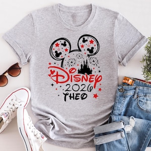 May include: A light gray t-shirt featuring a Disney-themed graphic. The design includes Mickey Mouse ears, stars, fireworks, a castle silhouette, and the text "Disney 2026 THEO" in red and black. The shirt is paired with jeans and white sneakers.