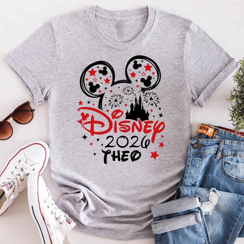 May include: A light gray t-shirt featuring a Disney-themed graphic. The design includes Mickey Mouse ears, stars, fireworks, a castle silhouette, and the text "Disney 2026 THEO" in red and black. The shirt is paired with jeans and white sneakers.