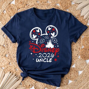 May include: Navy blue t-shirt with a Disney-themed design. The design includes Mickey Mouse ears, fireworks, a castle, and the text "Disney 2026 Uncle" in red and white. Stars and hearts complete the festive look.