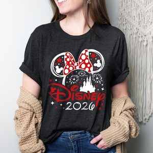 May include: Dark gray t-shirt featuring a Minnie Mouse bow with red polka dots and ears with red stars. The word "Disney" is in red script, with "2020" below. Fireworks and a castle silhouette complete the design.