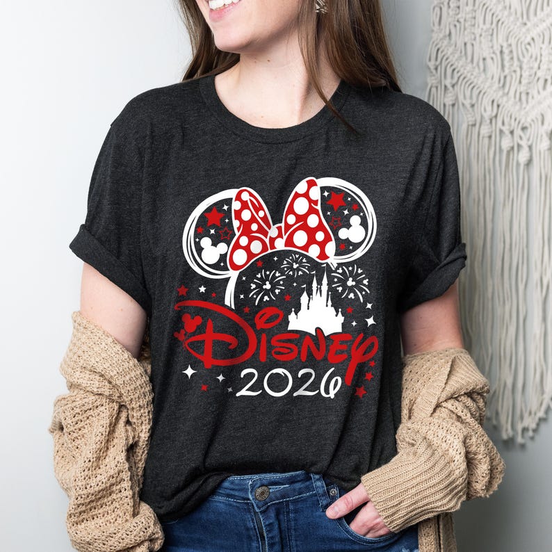 May include: Dark gray t-shirt featuring a Minnie Mouse bow with red polka dots and ears with red stars. The word "Disney" is in red script, with "2020" below. Fireworks and a castle silhouette complete the design.