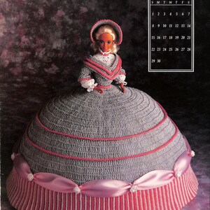 May include: A crocheted doll wearing a grey and pink dress with a white lace collar and a pink hat. The doll is standing on a grey background with a calendar for September in the background.