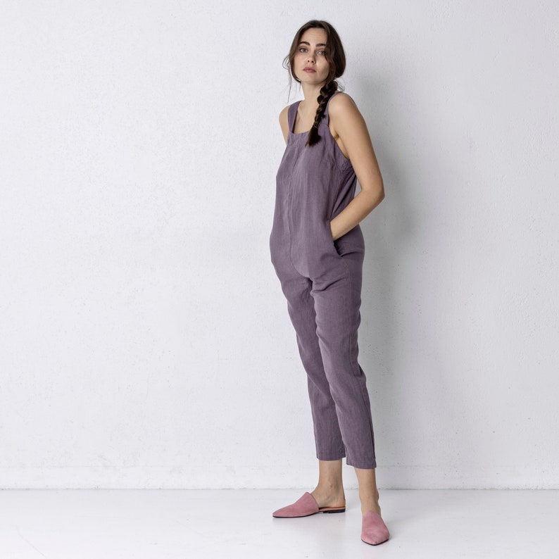 May include: A woman wearing a purple linen jumpsuit with a square neckline and thin straps. She is standing with her hands in her pockets and is wearing pink slip-on shoes.