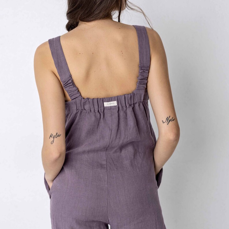 May include: A purple linen jumpsuit with adjustable straps. The jumpsuit has a relaxed fit and a slightly cropped leg. The model is wearing the jumpsuit with the straps adjusted to show off the back.