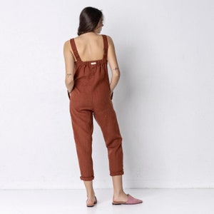 May include: A rust-coloured linen jumpsuit with thin straps and a relaxed fit. The jumpsuit has a wide leg and a slightly cropped length.