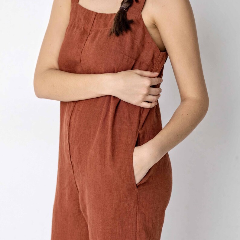 May include: A rust-coloured linen jumpsuit with a sleeveless design and two pockets. The jumpsuit has a relaxed fit and a straight leg.