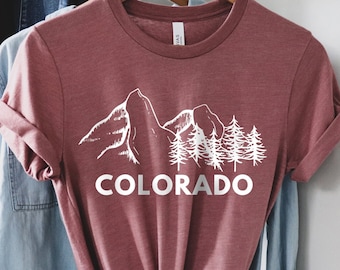 Op de afbeelding: Een maronkleurig T-shirt met een witte afbeelding van bergen en bomen. De tekst "COLORADO" is onder de afbeelding gedrukt.