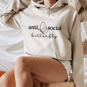Może przedstawiać: Beżowa bluza z kapturem z czarnym nadrukiem motyla i tekstem "anti-social butterfly" na przodzie.
