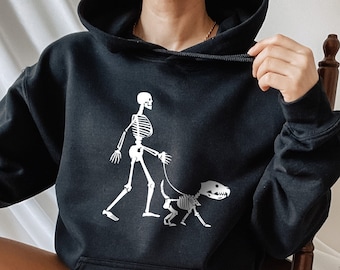May include: A black cropped hoodie with a white skeleton graphic walking a skeleton dog. The hoodie has a drawstring hood and a kangaroo pocket.
