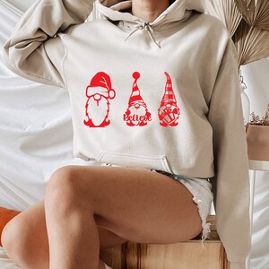 May include: A beige hoodie with a red graphic of three gnomes. The gnomes are wearing Santa hats and one is holding a gift. The text "Believe" is printed below the gnomes.