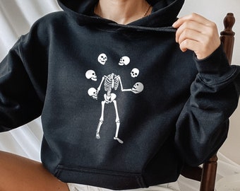 May include: A black hoodie with a white skeleton graphic juggling skulls. The hoodie has a drawstring hood and a kangaroo pocket.