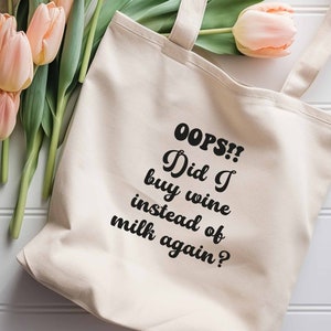 May include: A white canvas tote bag with black text that reads "OOPS!! Did I buy wine instead of milk again?" The bag is lying on a white wooden surface with a bouquet of pink tulips.