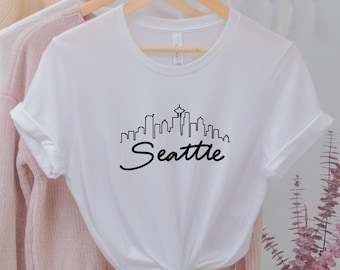 May include: White t-shirt with a black graphic of the Seattle skyline and the word "Seattle" in a cursive font.