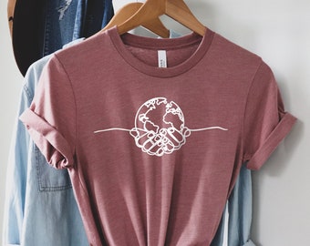 May include: A mauve t-shirt with a white graphic of hands holding a globe. The shirt is tied at the bottom.