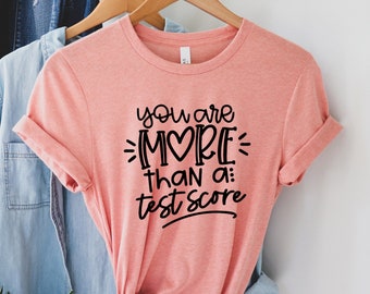 Könnte beinhalten: Ein pfirsichfarbenes T-Shirt mit einer schwarzen Grafik, auf der "You are more than a: test score" mit einem Herzen im Wort "more" steht.
