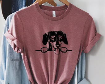 Könnte beinhalten: Ein mauvefarbenes T-Shirt mit einer schwarzen Silhouette eines Dackels, der über den oberen Rand schaut. Der Dackel hat die Zunge herausgestreckt.
