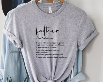 May include: Grey t-shirt with the word "father" and its definition printed in black text. The definition reads: "A man who loves, supports, guides, inspires and encourages his children. Who teaches by example. One who is patient, kind and understanding. Provider, protector, mentor, hero. A man who is admired and very loved."
