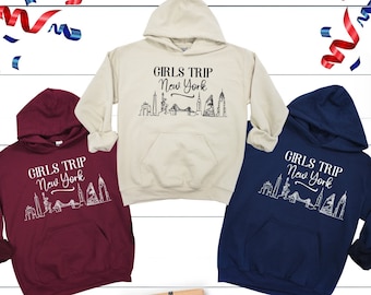 May include: Three hooded sweatshirts in burgundy, cream, and navy blue, each with the text "GIRLS TRIP New York" and a skyline graphic. The sweatshirts are displayed with gift boxes and confetti, suggesting a travel or gift theme.
