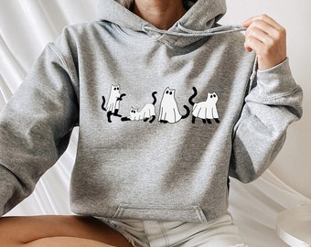 Puede incluir: Una sudadera gris con capucha con un gráfico de cinco gatos blancos vestidos de fantasmas. La sudadera la lleva puesta una persona sentada en un taburete de madera.