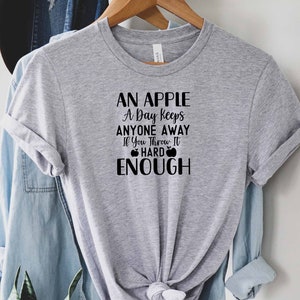 May include: Gray t-shirt with a black graphic that reads "An apple a day keeps anyone away if you throw it hard enough" with two apple icons.