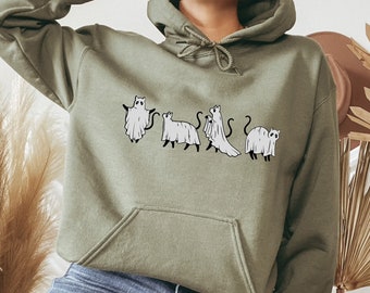 Puede incluir: Una sudadera con capucha verde con un gráfico de cinco gatos vestidos de fantasmas. Los gatos están en fila, con el primer gato mirando hacia la izquierda, el segundo gato mirando hacia la derecha, el tercer gato mirando hacia la izquierda, el cuarto gato mirando hacia la derecha y el quinto gato mirando hacia la izquierda. La sudadera la lleva puesta una persona sentada en un taburete de madera.