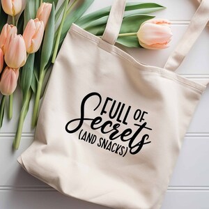 Puede incluir: Una bolsa de lona blanca con texto negro que dice "Full of Secrets (and Snacks)". La bolsa está sobre una superficie de madera blanca con un ramo de tulipanes rosas.