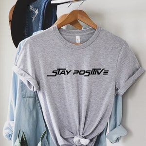 Puede incluir: Camiseta gris con texto negro que dice "STAY POSITIVE" en una fuente estilizada.