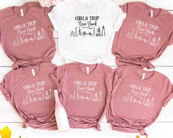May include: Five pink t-shirts with the text "Girls Trip New York" and a graphic of the New York City skyline in white. The shirts are arranged in a circle on a white surface.