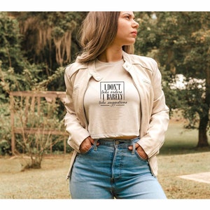 May include: A woman wearing a white crop top with the text "I DON'T take orders I BARELY take suggestions" in a black box. She is wearing a light brown leather jacket and blue jeans.