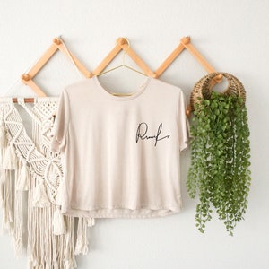 May include: A beige cropped t-shirt with the word "Remark" written in black cursive on the front. The shirt is hanging on a wooden hanger against a white wall.