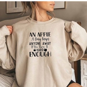 May include: A beige sweatshirt with a black graphic that reads "An apple a day keeps anyone away if you throw it hard enough" with two apple illustrations.