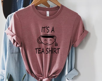 May include: A mauve t-shirt with a black graphic of a teacup with a smiling face and the text "It's a Tea Shirt".