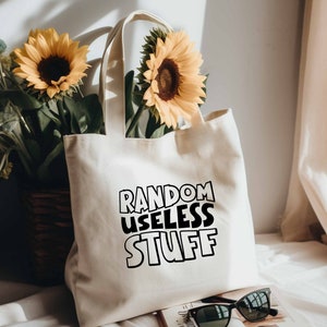 May include: A white canvas tote bag with black text that reads "RANDOM USELESS STUFF". The bag is sitting on a white surface with a book and sunglasses next to it. There are sunflowers in the background.