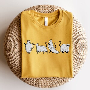 May include: A yellow t-shirt with a graphic of four cats dressed as ghosts. The cats are in a line, facing forward, and each has a white ghost sheet draped over their body. The text "Canvas" is visible on the shirt's tag.