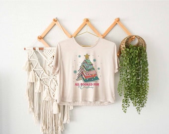 May include: A beige t-shirt with a graphic of a Christmas tree made of stacked books. The text "All Booked for Christmas" is printed below the tree.