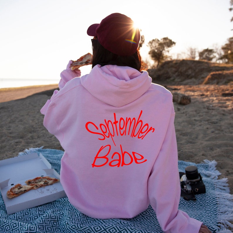 Puede incluir: Una sudadera con capucha rosa con el texto "September Babe" en letras rojas en la parte posterior.