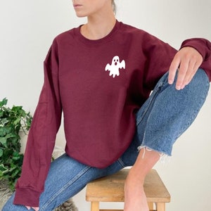 May include: A maroon sweatshirt with a white ghost graphic on the front. The sweatshirt is worn by a person sitting on a wooden stool.