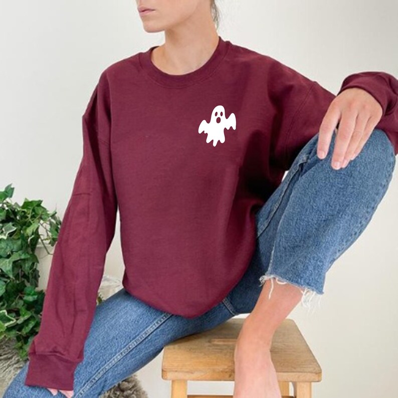 May include: A maroon sweatshirt with a white ghost graphic on the front. The sweatshirt is worn by a person sitting on a wooden stool.