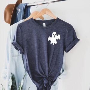 以下が含まれることがあります： 前面に白い幽霊のグラフィックが描かれた濃い青色のTシャツ。幽霊は驚いた表情をして、白いシーツを着ています。