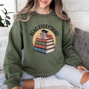 May include: Olive green sweatshirt with the text "READ EVERYTHING" in a circular design. The graphic features a cartoon worm wearing a graduation cap and glasses, sitting atop a stack of colorful books.