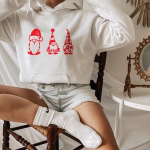 May include: White hoodie with a red graphic of three gnomes wearing Santa hats. The gnome in the middle has the word "Believe" written on its hat.