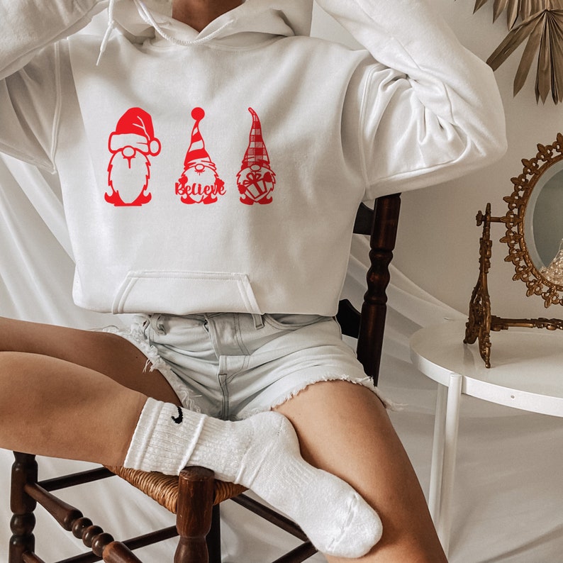 May include: White hoodie with a red graphic of three gnomes wearing Santa hats. The gnome in the middle has the word "Believe" written on its hat.