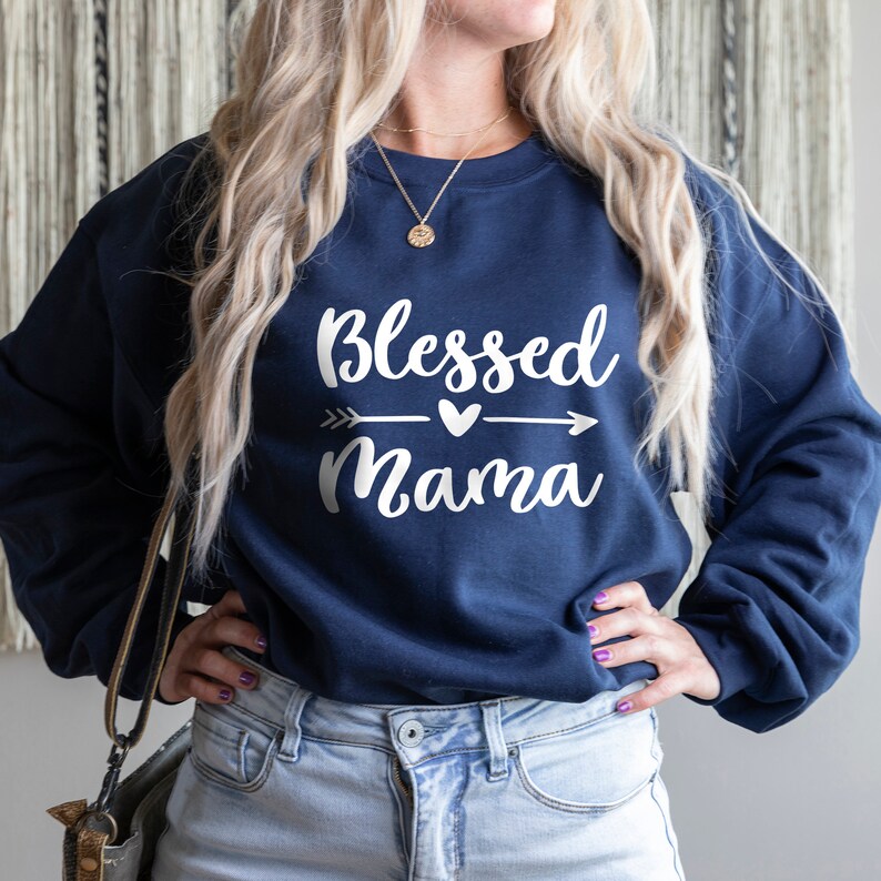 Puede incluir: Una sudadera azul marino con el texto "Blessed Mama" en letras blancas. El texto est&aacute; acompa&ntilde;ado por un peque&ntilde;o coraz&oacute;n y una flecha que apunta hacia la derecha.